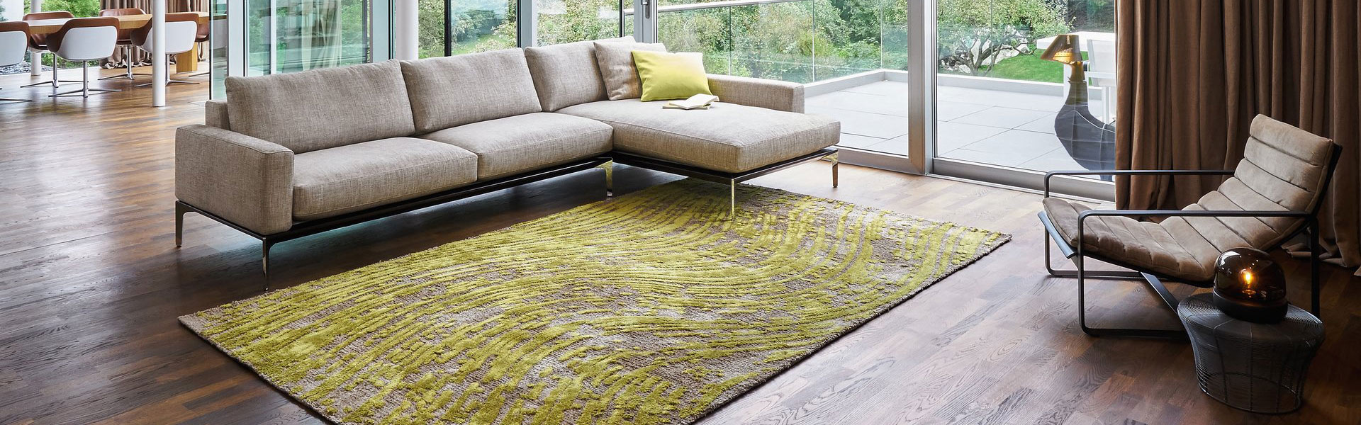 Modernes Wohnzimmer mit beigem Sofa, grünem Teppich und Glaswand mit Gartenblick.