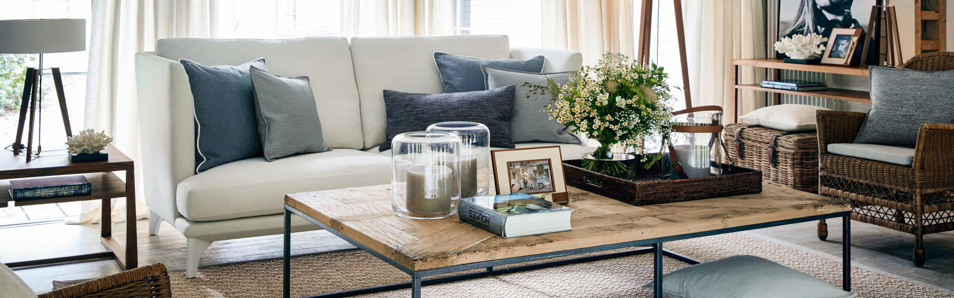 Helles Wohnzimmer mit weißem Sofa, grauen Kissen, Holztisch und grünem Blumenschmuck.